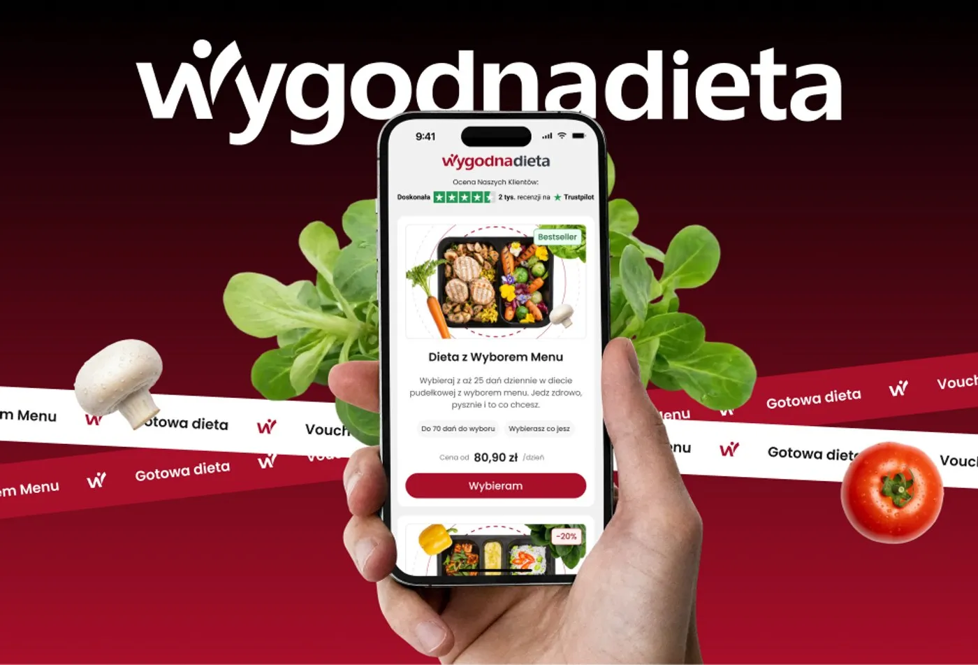 WygodnaDieta - telefon w dłoni z ekranem głównym aplikacji: ocena „Doskonała" na Trustpilocie (2 tys. recenzji) i karta diety „Dieta z Wyborem Menu" z ceną od 80,90 zł/dzień oraz przyciskiem „Wybieram", na tle czerwonego gradientu z warzywami i wstęgą z nazwami produktów.