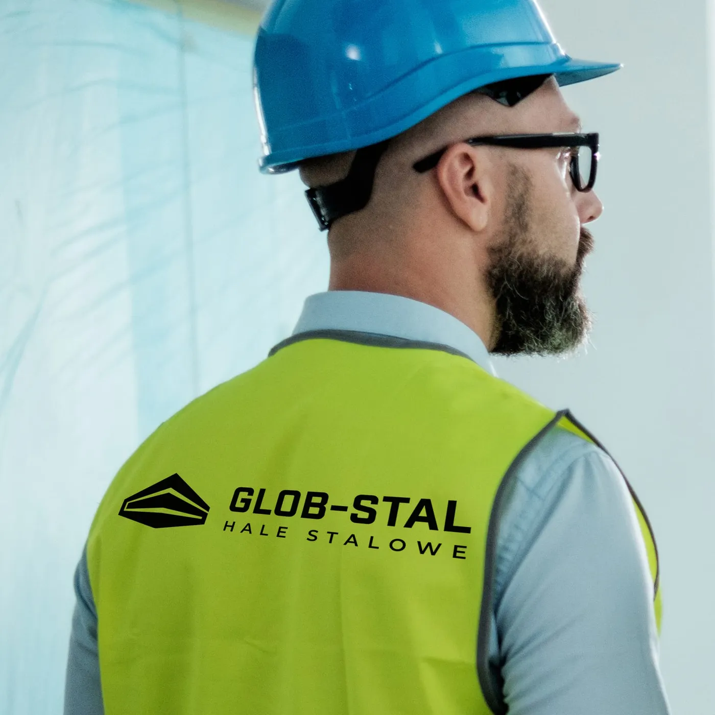 Glob-Stal - pracownik w kasku i żółtej kamizelce odblaskowej z czarnym logo Glob-Stal i napisem „Hale stalowe" na plecach, na tle budowy.