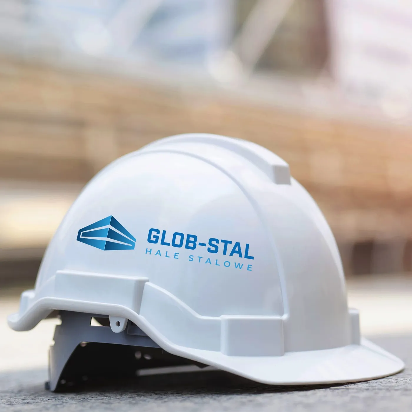 Glob-Stal - biały kask ochronny z niebieskim logo w kształcie diamentu i logotypem „Glob-Stal - Hale stalowe", na tle placu budowy.