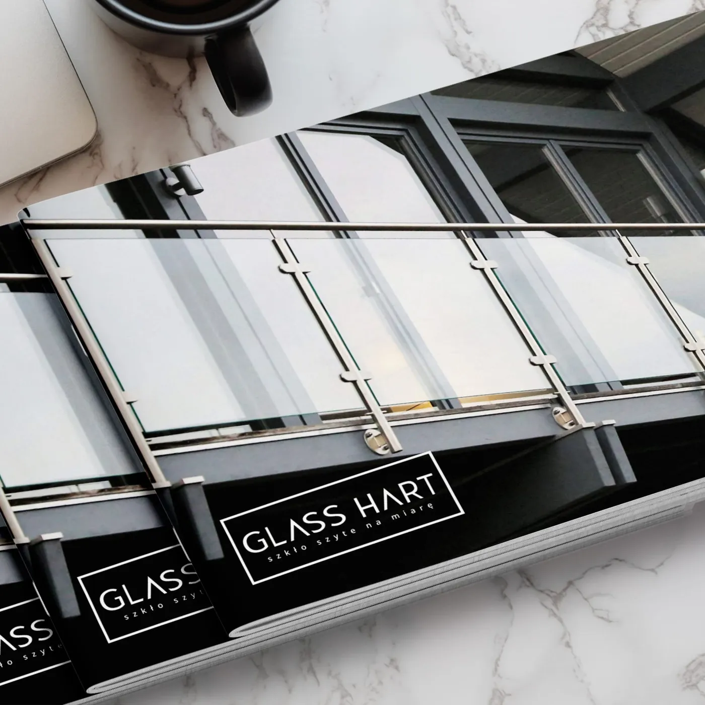 Glass Hart - zamknięty katalog leżący na fotografii szklanej balustrady balkonowej, z czarną okładką i logotypem „Glass Hart - szkło szyte na miarę".
