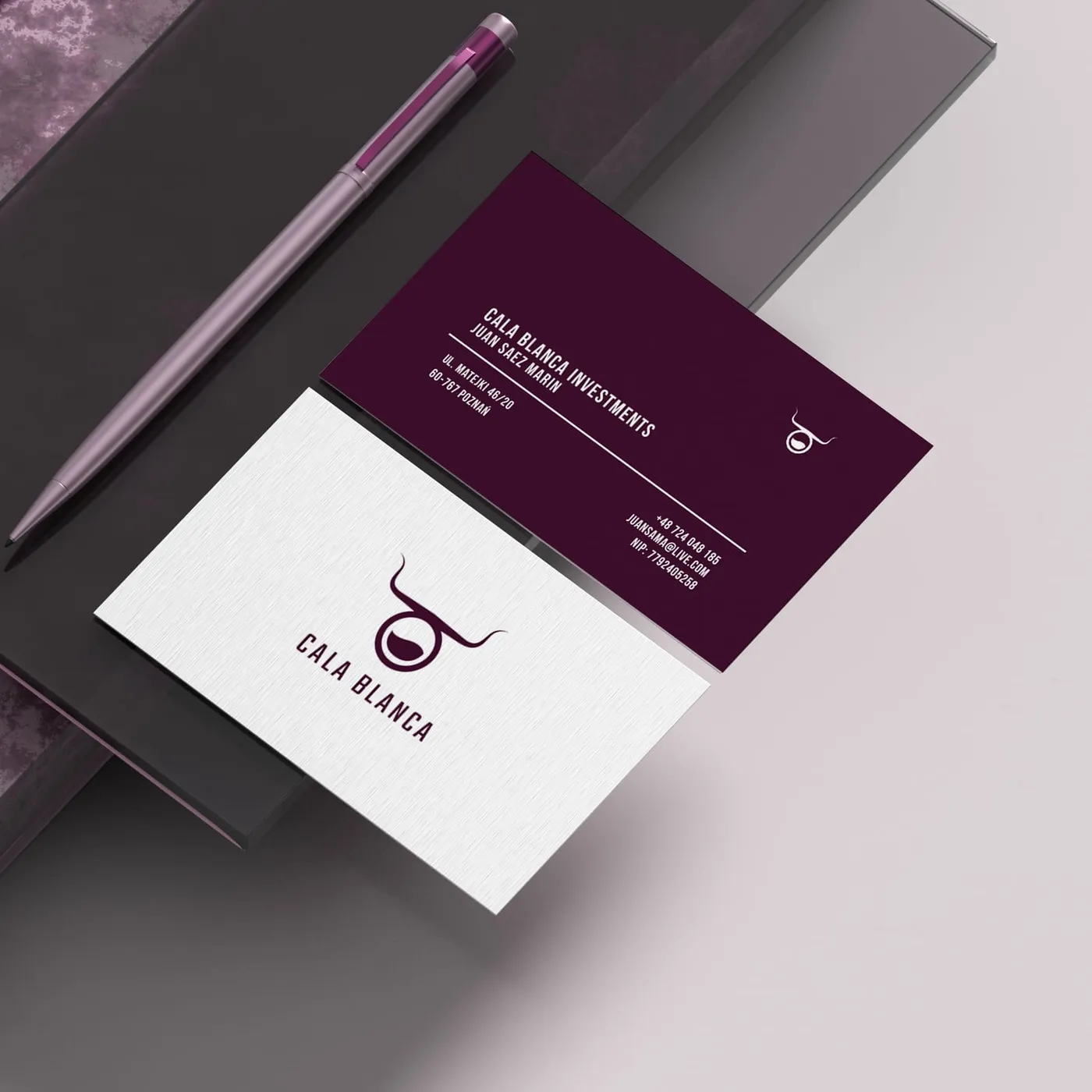 Para wizytówek Cala Blanca na ciemnym stole obok pióra - biała wizytówka z logo byczej głowy oraz bordowa wizytówka z danymi firmy „Cala Blanca Investments".