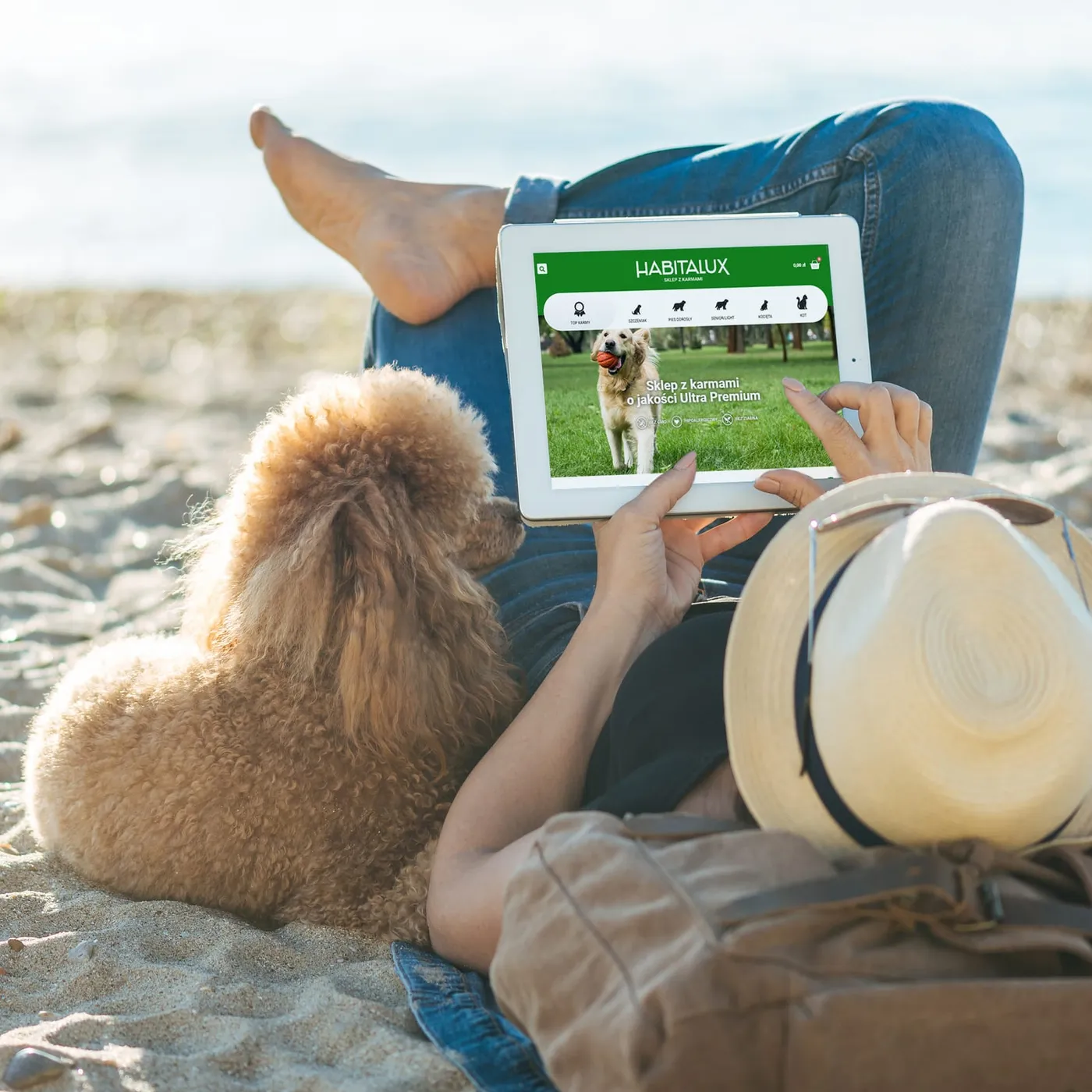 Habitalux - tablet z otwartą stroną główną sklepu internetowego („Sklep z karmami o jakości Ultra Premium") trzymany przez osobę siedzącą na plaży z psem.