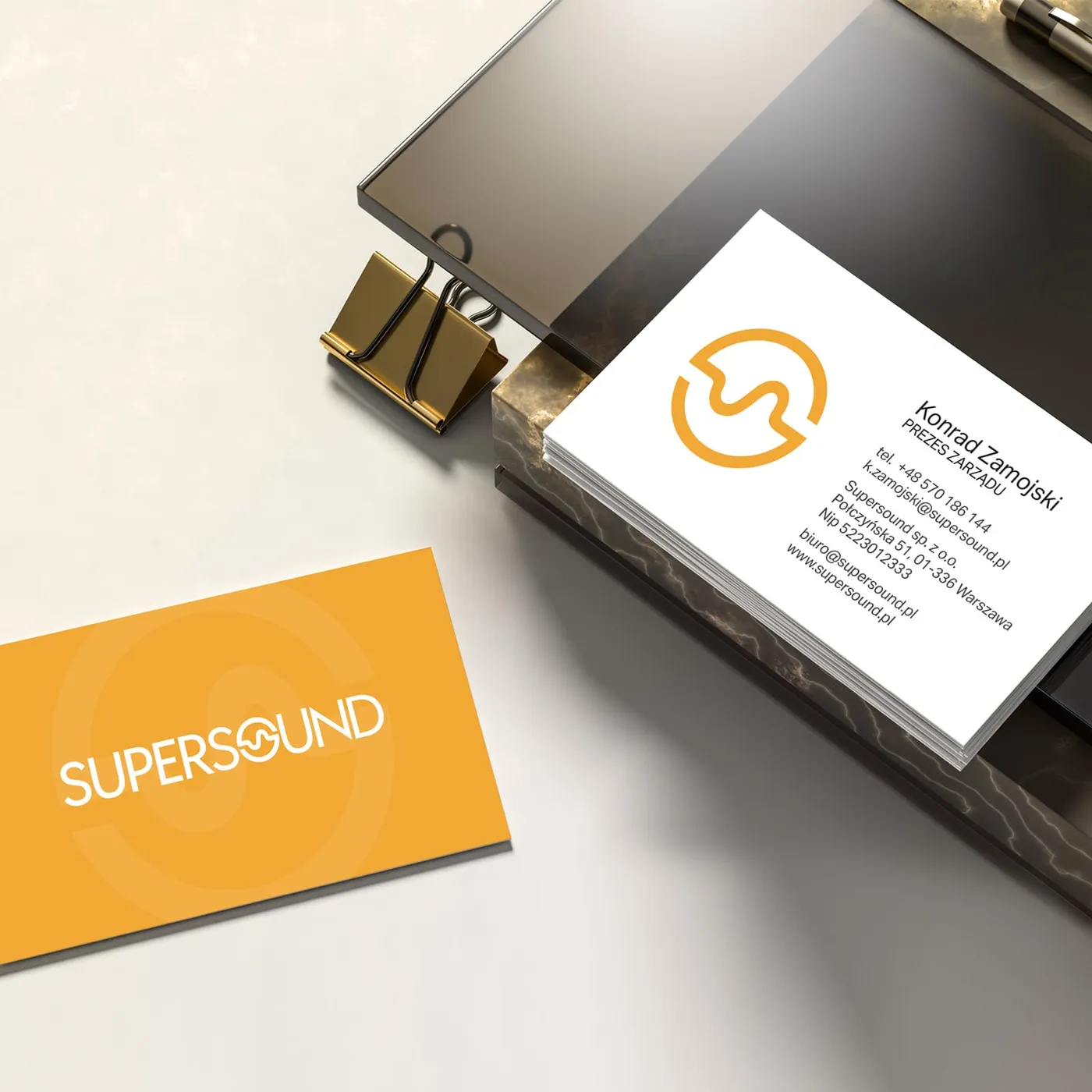 Supersound - wizytówka Konrada Zamojskiego, Prezesa Zarządu, z danymi kontaktowymi i adresem spółki, oparta o stos wizytówek spiętych złotym klipsem.