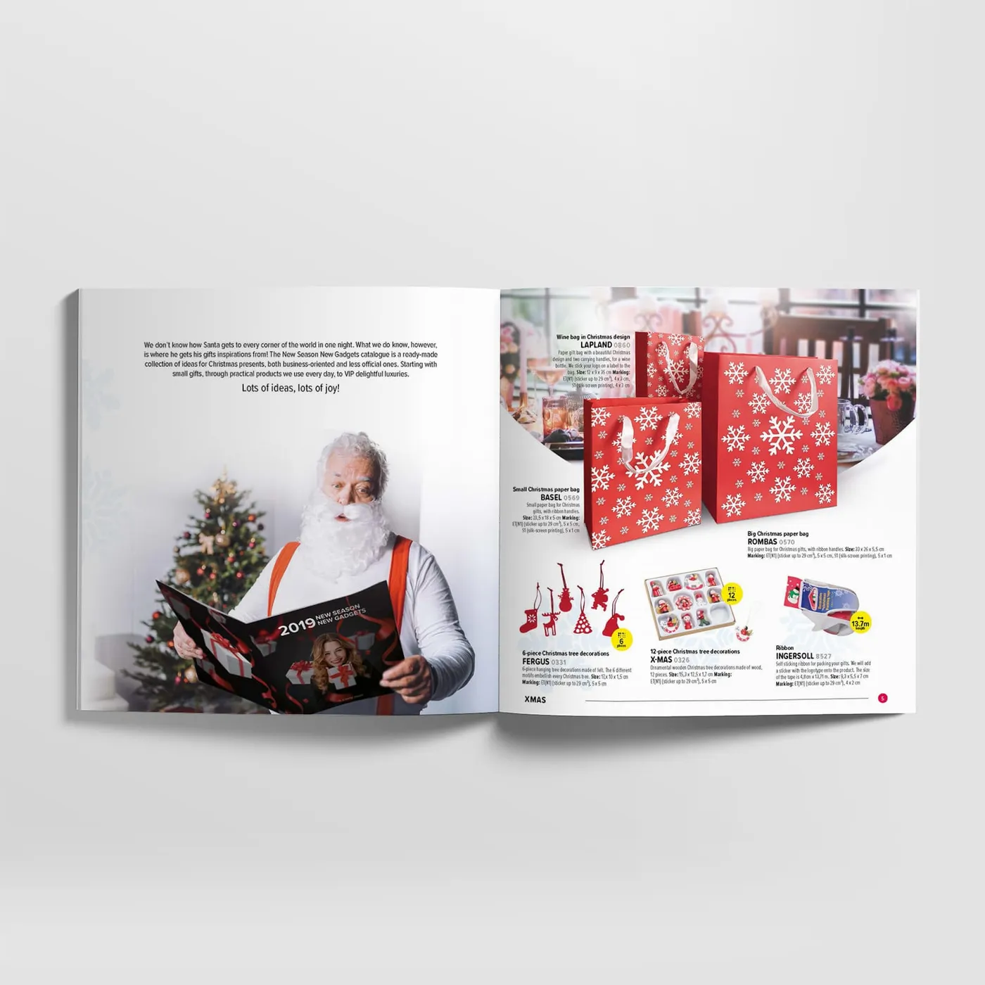 Macma - Święty Mikołaj czytający anglojęzyczny katalog „2019 New Season New Gadgets" z czerwoną okładką, obok choinki; na sąsiedniej stronie czerwone torby prezentowe we wzór śnieżynek.